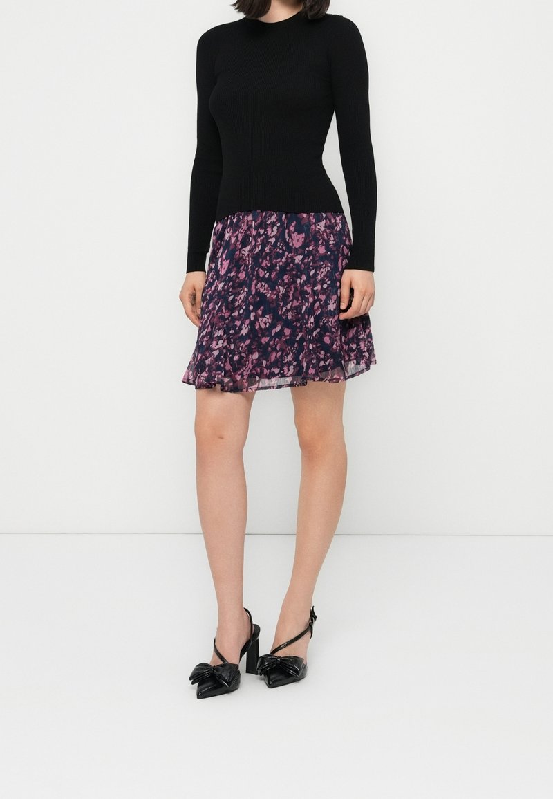 Femme portant un haut noir à manches longues, une jupe fleurie violette et des chaussures à talons noirs avec des nœuds, debout sur fond blanc.
