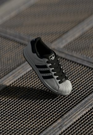 Grå Adidas sneaker med sorte striber og snørebånd, der svæver over en tekstureret metalristet overflade.