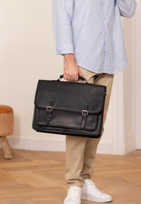 Black leather briefcase with dual buckle closures, flat design, sturdy handle, and contrasting stitching, held by a person.