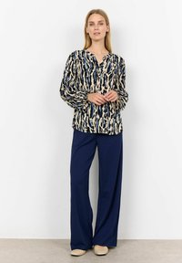 Blusa fluida in crema con stampa astratta blu scuro e nera, dotata di maniche lunghe e colletto con bottoni, abbinata a pantaloni navy.