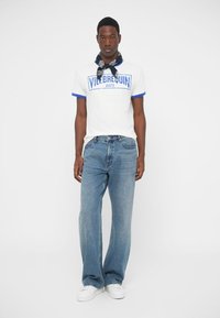 Bela bombažna majica s plavimi detajli in logotipom, v kombinaciji z modrimi jeans hlačami. Model nosi bele športne copate in temno modro ruto.