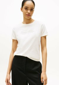 Camiseta de algodón blanca con el logo bordado de "Tommy Hilfiger" en cursiva. Manga corta y escote redondo, combinada con pantalones negros.