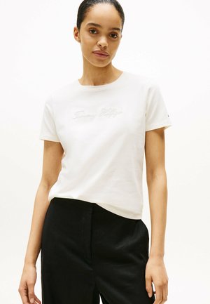 T-shirt en coton blanc avec logo brodé "Tommy Hilfiger" en cursive. Manches courtes et encolure ronde, associé à un pantalon noir.