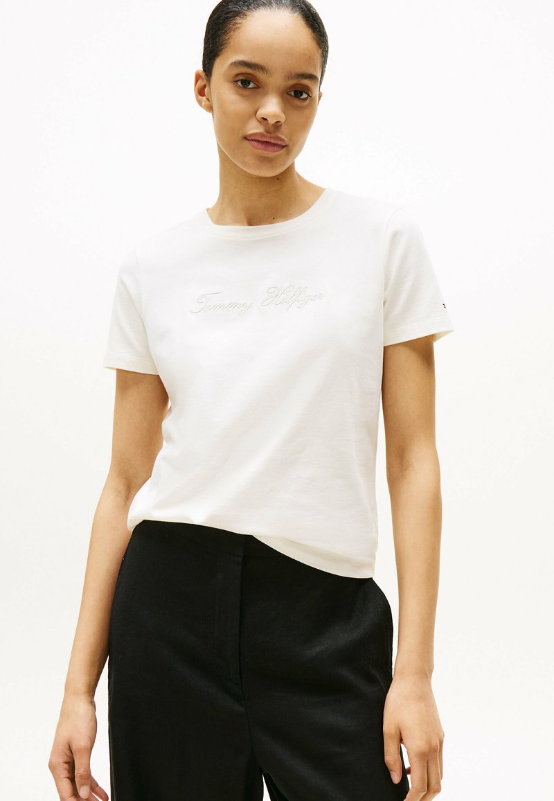 Camiseta de algodón blanca con el logo bordado de "Tommy Hilfiger" en cursiva. Manga corta y escote redondo, combinada con pantalones negros.