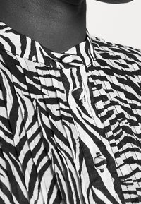 Košile s černobílým zebřím potiskem, zaobleným límečkem, zapínáním na knoflíky a vertikálními sklady pro texturu. Detailní pohled na horní část oděvu.