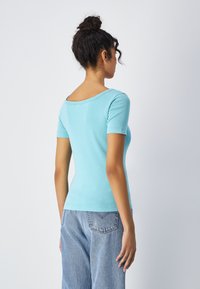 Jasnoniebieska dopasowana koszulka z krótkim rękawem, okrągłym dekoltem i gładką fakturą. Noszona z luźnymi niebieskimi dżinsami, widok z tyłu.