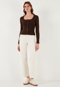 Bruine geribbelde cardigan met een gegolfde halslijn, knopen aan de voorkant en lange mouwen, gecombineerd met witte wijde jeans en beige sneakers.