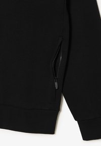 Primo piano di un indumento nero a maniche lunghe che mostra una tasca con zip parzialmente aperta sul lato anteriore e orlo e polsini a coste.