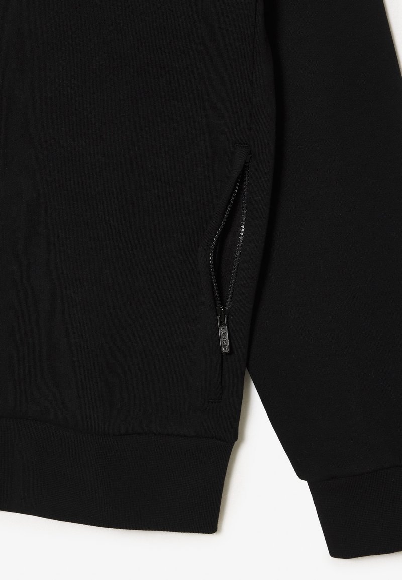 Primo piano di un indumento nero a maniche lunghe che mostra una tasca con zip parzialmente aperta sul lato anteriore e orlo e polsini a coste.