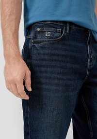 Granatowe dżinsy o prostym kroju, z delikatną teksturowaną powierzchnią, przeszyciami dekoracyjnymi oraz małym logo marki na tylnej kieszeni.