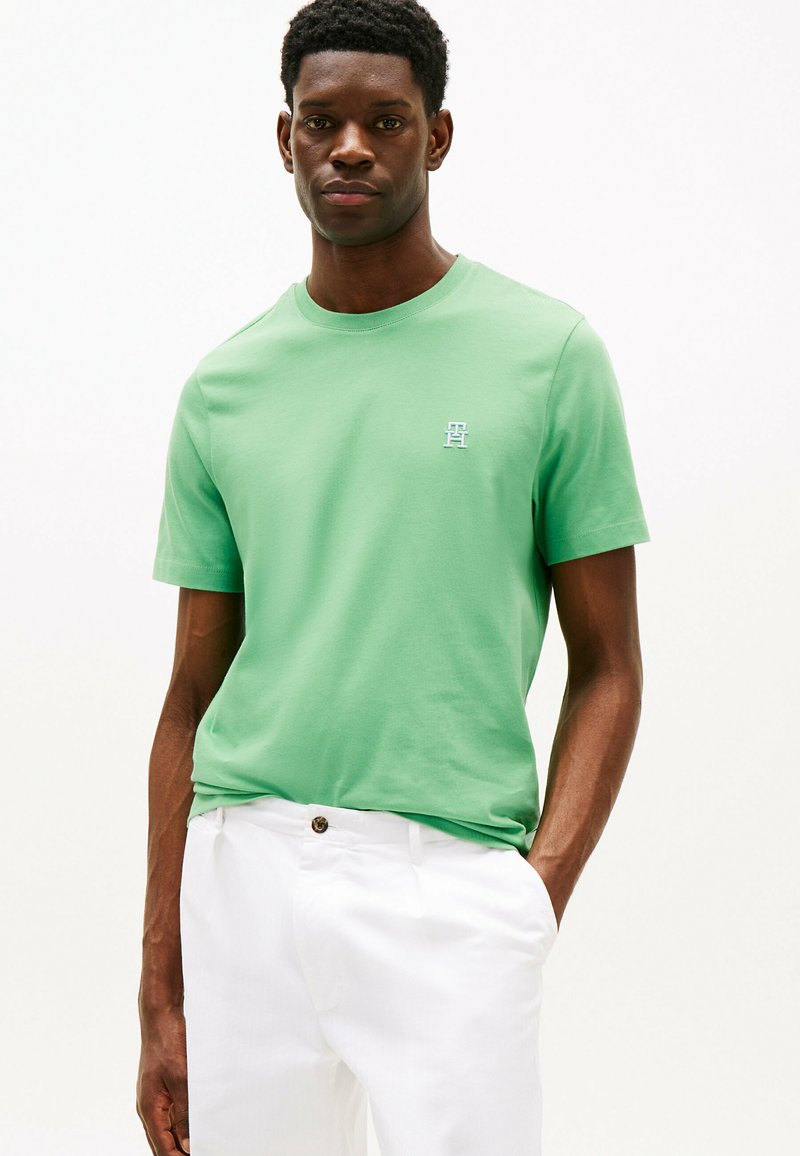T-shirt verde in cotone con collo tondo e maniche corte, caratterizzato da un piccolo logo azzurro sul petto, abbinato a pantaloni bianchi.