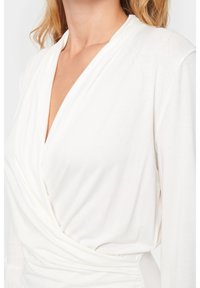 White wrap top with long sleeves, made from soft fabric. Features a deep V-neckline and gathered detailing at the waist.