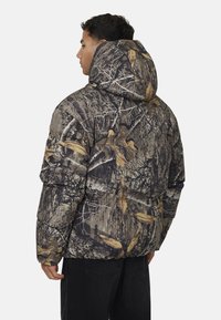 Kurtka puchowa w kamuflażowy wzór w ziemistych odcieniach, z kapturem, pikowaną strukturą i warstwową teksturą dla lepszej izolacji.