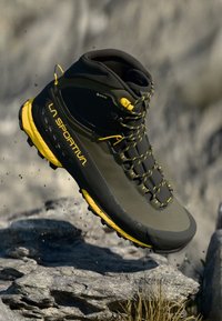 Čierno-žltá turistická obuv La Sportiva vo vzduchu nad kamenistým terénom, predvádzajúca robustnú podrážku a GORE-TEX svrchný materiál pre vonkajšie použitie.