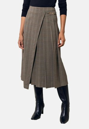 Mujer con falda cruzada a cuadros marrón hasta la rodilla con detalle de hebilla, camiseta negra de manga larga y botas de cuero negras hasta la rodilla.