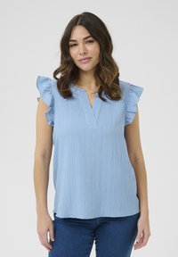 Top corto a maniche corte di colore azzurro con dettagli a ruches, tessuto strutturato e design a scollo a V. Indossato su jeans aderenti blu.