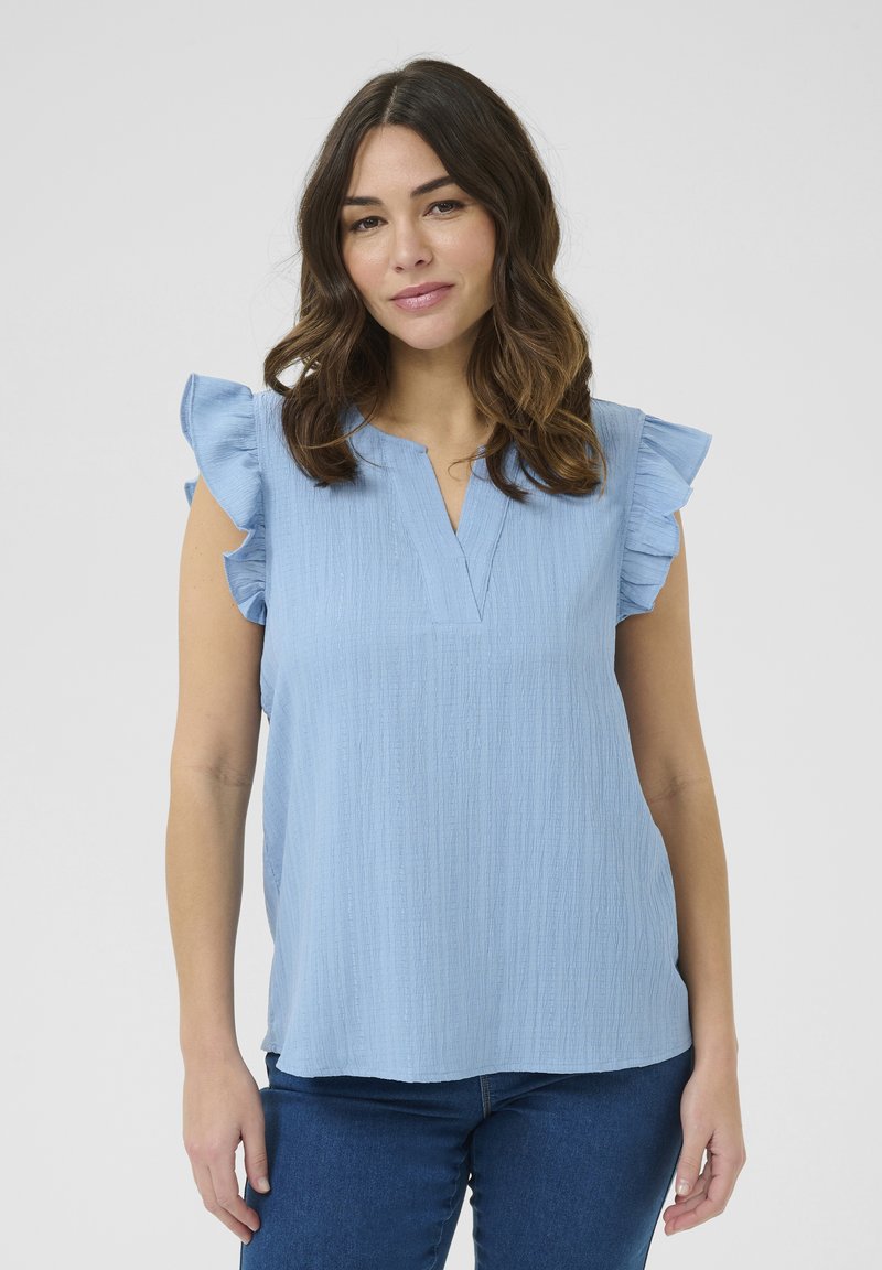 Top corto a maniche corte di colore azzurro con dettagli a ruches, tessuto strutturato e design a scollo a V. Indossato su jeans aderenti blu.