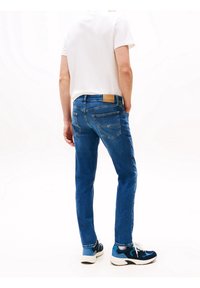 Niebieskie wąskie dżinsy z denimu, z naszywką skórzaną na pasie i tylnych kieszeniach. Noszone z białym T-shirtem i sneakersami.