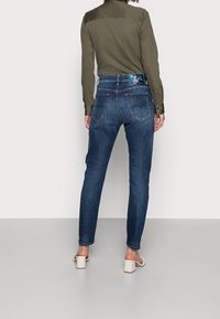 Jeans de mezclilla azul oscuro con ajuste tapered, que presentan un diseño floral en la cintura y bolsillos traseros visibles. Combinados con una camisa verde.