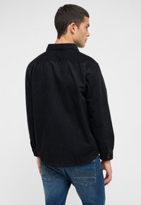 Schwarzes Cordhemd mit einem Button-Down-Kragen und langen Ärmeln, mit einer strukturierten Oberfläche und einem gerade geschnittenen Rücken. Kombiniert mit blauen Jeans.