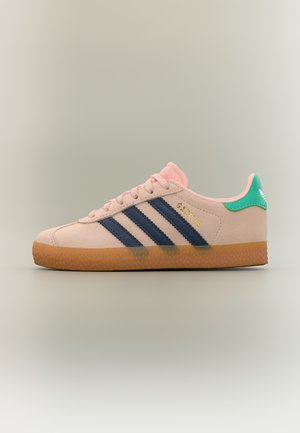 Svetloružičasta Adidas Gazelle tenisica s tamnoplavim prugama, zelenim jezičkom na peti, gumiranim potplatom i zlatnim natpisom "GAZELLE" sa strane.