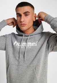 Szara bluza z kapturem, wykonana z miękkiego materiału, z przednią kieszenią i białym logo z napisem "hummel" oraz "SPORTSWEAR SINCE 1923".