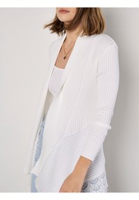 Žena na sobě má bílou žebrovanou cardigan over bílým tílkem, doplněnou světle modrými džíny a zlatým náhrdelníkem ve tvaru srdce.