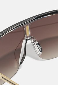 Γυαλιά ηλίου με φακούς καφέ gradient, γυαλιστερό μαύρο πλαίσιο και ασημένιες λεπτομέρειες; διαθέτουν μια διαφανή γέφυρα με χρυσή λεπτομέρεια.