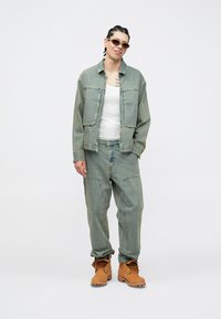 Gaiši zilas džinsa jaciņas un bikses, kombinētas ar baltu topu. Brūnas mežģīņu zābaki. Aksesuāri ietver saulesbrilles un redzamus tetovējumus.