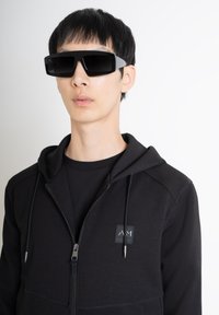 Gafas de sol con un grueso marco negro y acentos grises. Llevadas con una sudadera negra con cremallera que presenta un parche de logo y tejido texturizado.