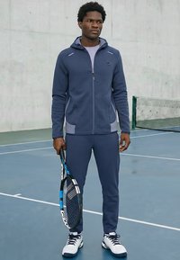 Homme debout sur un court de tennis tenant une raquette de tennis, vêtu d'un survêtement bleu marine et de chaussures de sport blanches.
