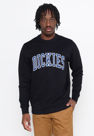 Man with black braided hair wears black sweatshirt with blue and white "DICKIES" text, and light brown pants, standing against white background.