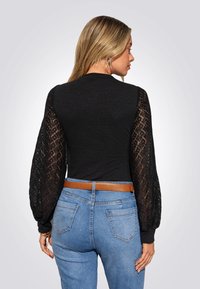 Schwarzer Body mit transparenten Spitzen Puffärmeln; hoher Kragen, figurbetontes Design und ein brauner Gürtel über hellblauen Jeans.