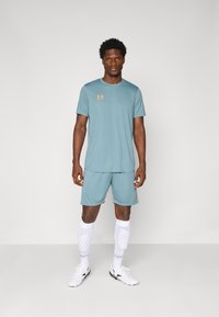 Athletic outfit featuring a short-sleeve teal shirt and matching shorts. White leg sleeves with dotted pattern, and white shoes. Orange logo detail.