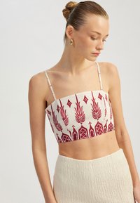 Kort top van crèmekleurige stof met ingewikkelde rode geometrische en bloemmotieven, voorzien van dunne, verstelbare banden.