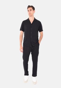 Camicia nera a maniche corte con tasca, abbinata a pantaloni neri e scarpe da ginnastica bianche. La camicia presenta un design con bottoni e una vestibilità comoda.