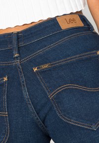 Dark blue denim jeans with a slim fit, featuring orange stitching and a brown leather Lee logo patch on the waistband. Two back pockets visible.