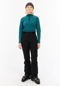 Femme portant un haut à manches longues teal, un pantalon de ski noir, des chaussures de ski noires et un bonnet gris foncé, se tenant sur un fond blanc.