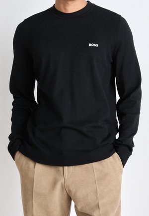 Mann trägt einen schwarzen Langarmpullover mit "BOSS"-Logo auf der Brust und beige Hose, Hände in den Taschen, vor einfachem Hintergrund.