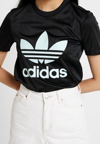 Musta adidas T-paita, jossa on iso vaaleansininen logo ja kolme valkoista raitaa rinnassa. Kangas näyttää sileältä ja kiiltävältä.