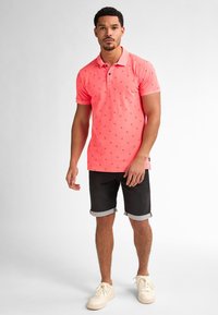 Ljusrosa polo med krage och små blå mönster, paired med svarta shorts som är uppvikt vid fållen och beige sneakers.