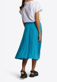 Falda midi plisada en azul brillante, hecha de tejido ligero. Presenta una silueta fluida y se combina con una blusa blanca de manga corta y sandalias oscuras.