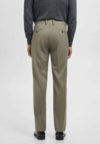 Mann trägt beigefarbene maßgeschneiderte Hose, dunkelgraues langärmeliges Hemd eingesteckt und schwarze Lederschuhe, steht mit dem Rücken zur Kamera vor einem einfarbigen Hintergrund.