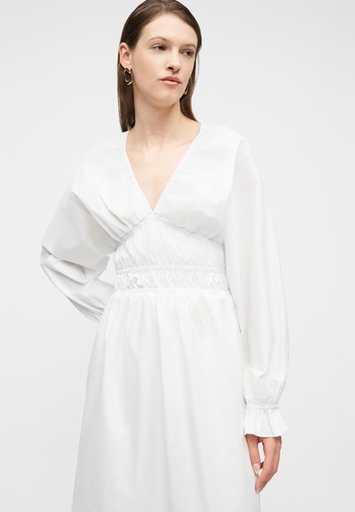 Γυναίκα που φοράει λευκό φόρεμα με μακριά μανίκια, βαθύ V στο ντεκολτέ και σφηκοφωλιά στη μέση, στέκεται μπροστά σε απλό φωτεινό φόντο.