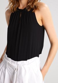 Svart halterneck-top i mjukt tyg, med en nyckelhålsdetalj vid halsen och ärmlös design, kombinerad med vita högmidjade byxor.