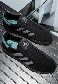 Baskets Adidas noires avec des bandes argentées et des logos Mercedes AMG sur les languettes, posées sur une surface métallique avec des reflets architecturaux.
