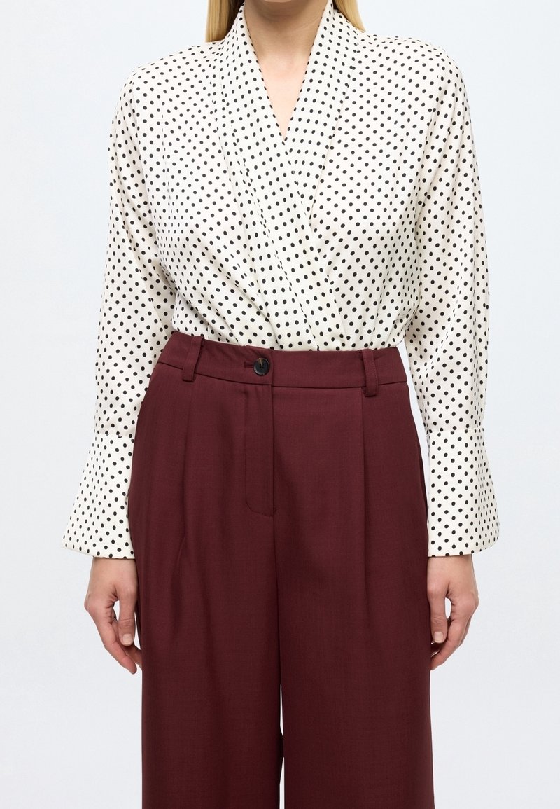Femme portant un chemisier blanc à manches longues à pois noirs, rentré dans un pantalon large bordeaux taille haute.