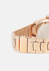 Rose gold metalurisk ur med et struktureret, sammenflettet armbånd og en poleret lås. Har et enkelt rundt urkasse og en glat bagside.