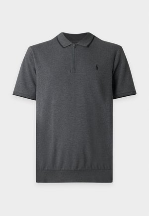 Homme portant un polo gris foncé à manches courtes, un pantalon cargo beige et des baskets blanches, debout avec une main dans la poche.
