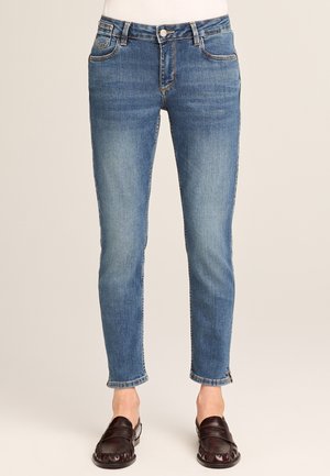 Frau trägt eng anliegende blaue Jeans aus Denim und dunkelbraune Slip-On-Loafer, steht vor einem schlichten beige Hintergrund.